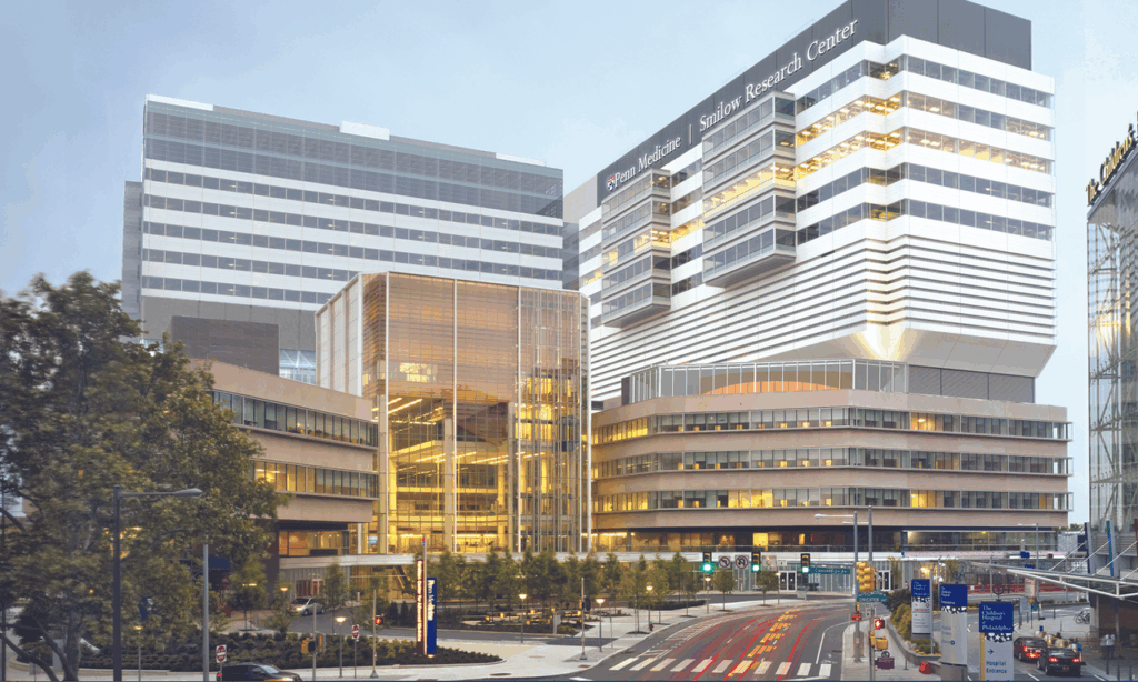 A modern, multi-story medical and research center with glass and metal facades, brightly lit interiors, and surrounding trees. A street with crosswalks and traffic signs runs in front of the building.