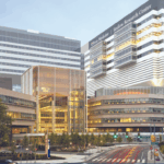 A modern, multi-story medical and research center with glass and metal facades, brightly lit interiors, and surrounding trees. A street with crosswalks and traffic signs runs in front of the building.