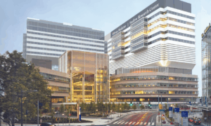 A modern, multi-story medical and research center with glass and metal facades, brightly lit interiors, and surrounding trees. A street with crosswalks and traffic signs runs in front of the building.