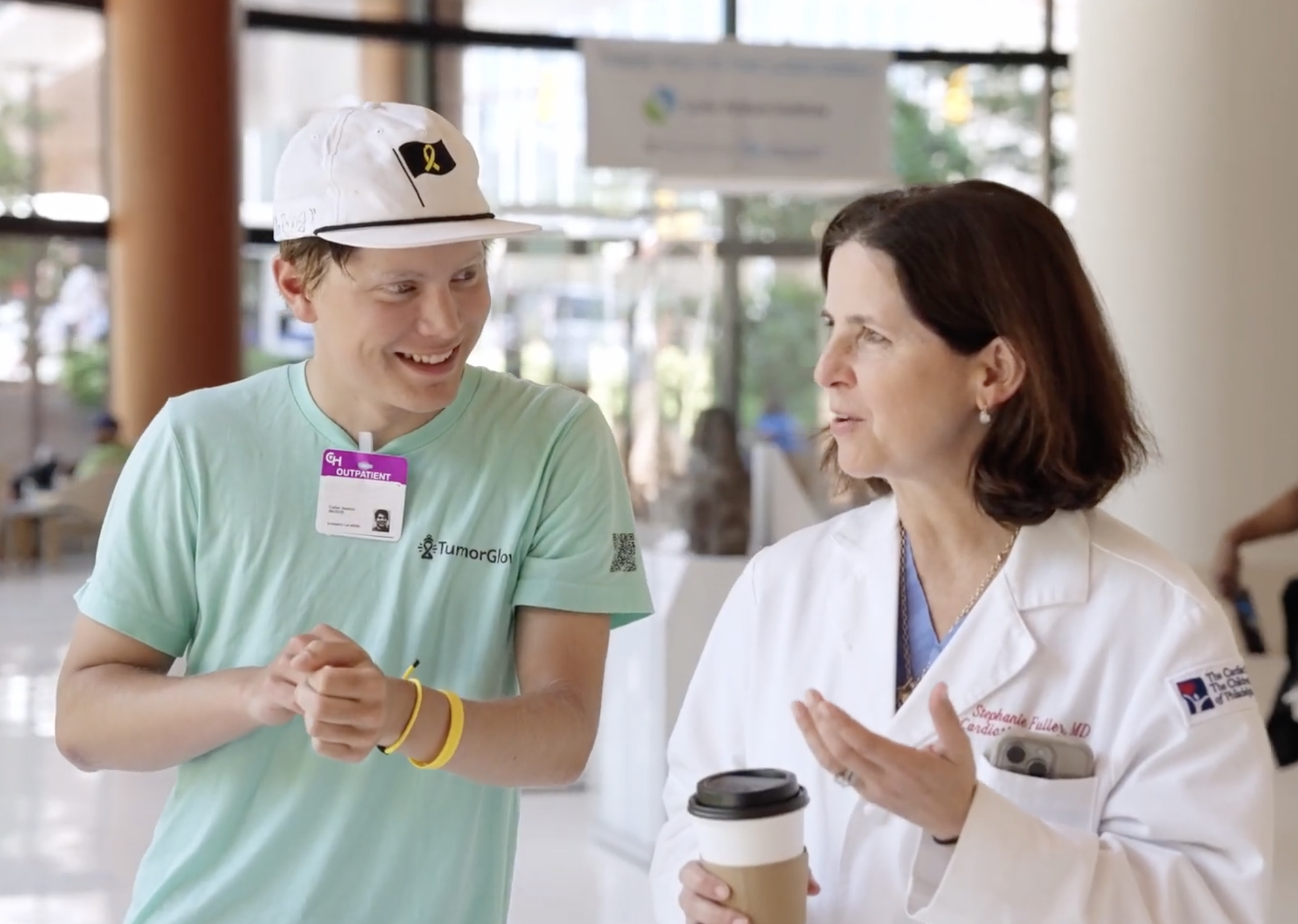 A young man wearing a white cap and light green shirt smiles while talking with a woman in a white lab coat holding a coffee cup, as they walk together in a bright, modern building.