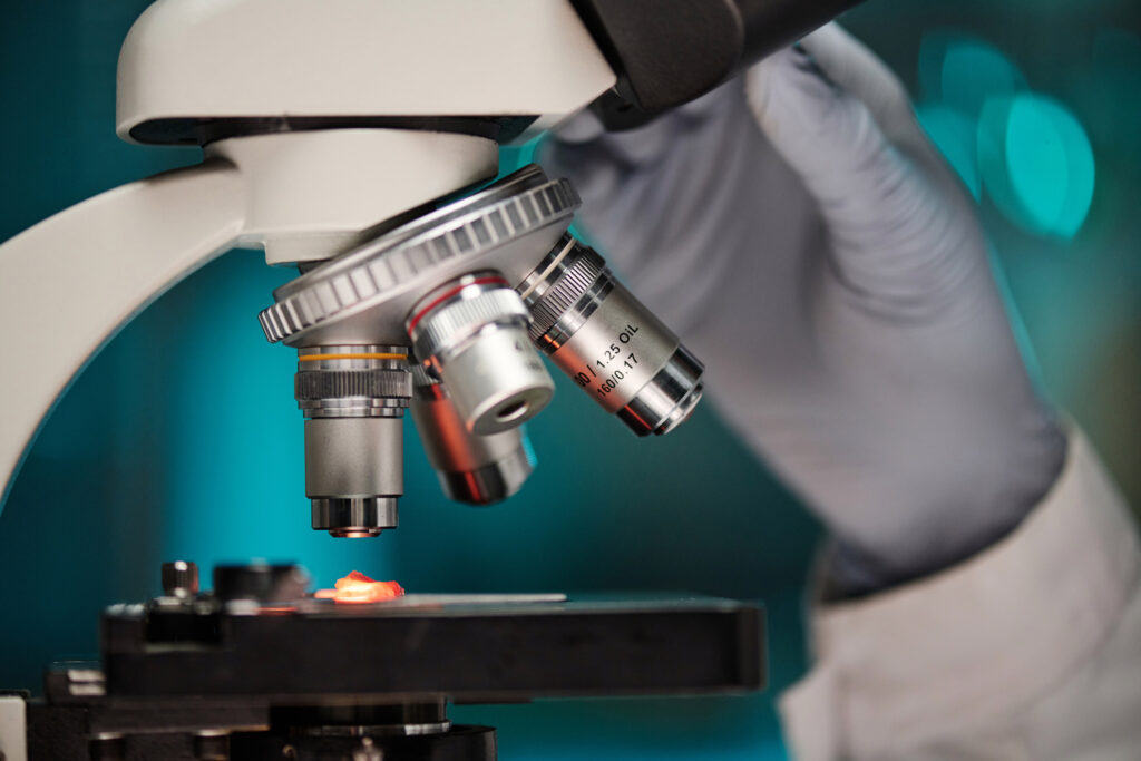 Close-up of a gloved hand adjusting the focus knobs on a laboratory microscope, with lens details visible and a specimen slide illuminated on the stage.