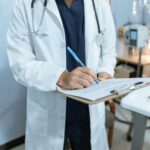 A doctor in a white coat and stethoscope writes on a clipboard while standing in a medical office or hospital room. Medical equipment and a bed are visible in the background.