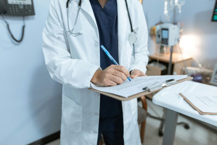 A doctor in a white coat and stethoscope writes on a clipboard while standing in a medical office or hospital room. Medical equipment and a bed are visible in the background.