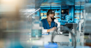 A scientist in blue scrubs looks into a microscope in a modern laboratory, surrounded by glassware, test tubes, and laboratory equipment with shelves of supplies in the background.