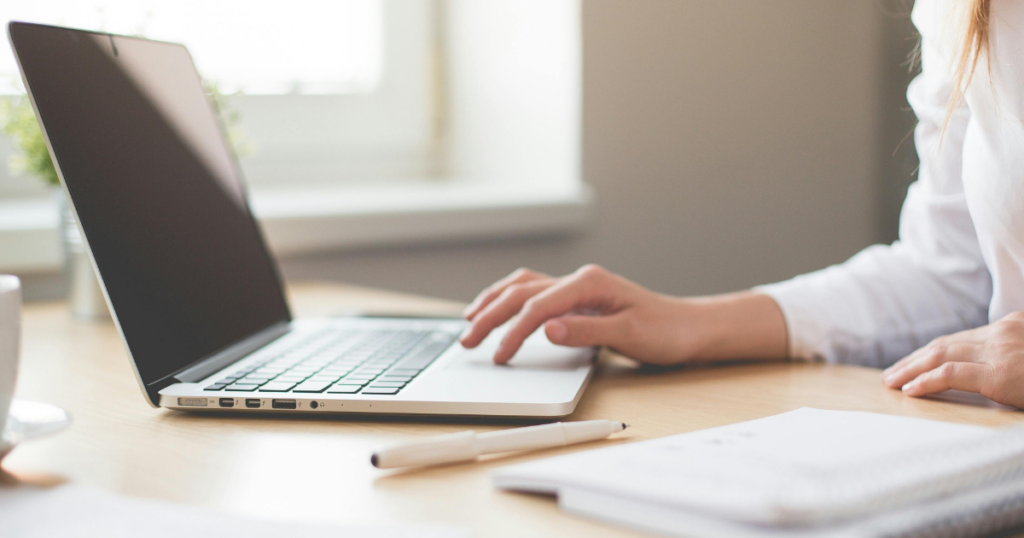 A person works on a laptop at a desk near a window, with a notebook and pen nearby. The scene is bright and airy, suggesting a calm and productive workspace.