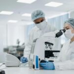 Two scientists in protective clothing and masks work in a laboratory; one is looking through a microscope while the other stands beside, observing and discussing the research.