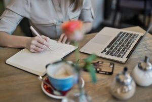 A person writes in a notebook next to an open laptop, a smartphone, a teacup with a saucer, and sugar bowls on a wooden table. A pink flower in a vase is blurred in the foreground.
