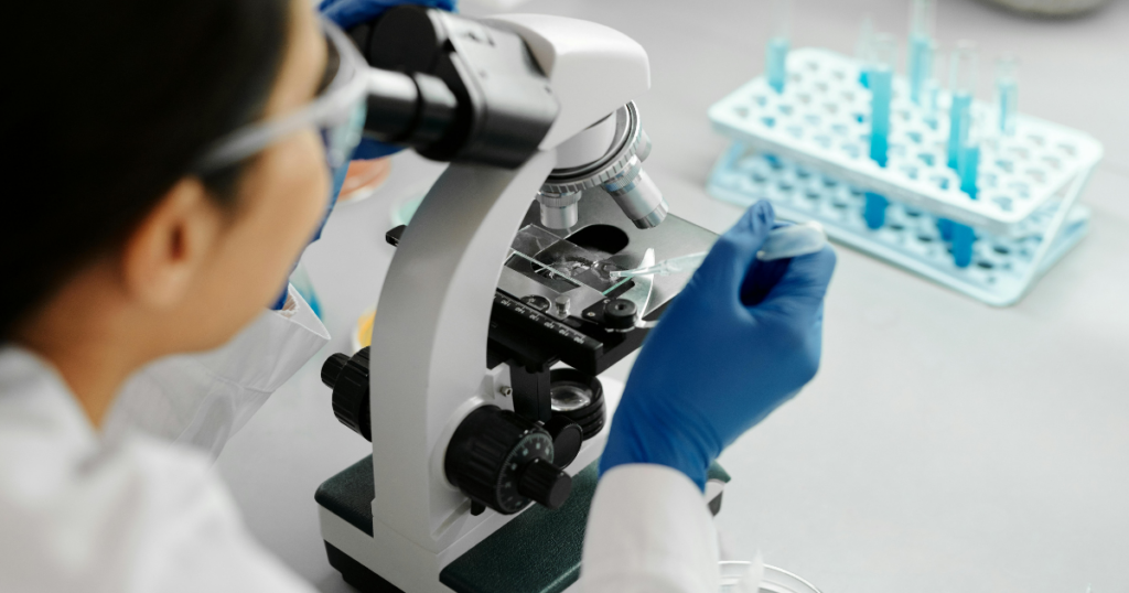 A scientist wearing blue gloves and safety goggles uses a pipette to place a sample on a glass slide under a microscope, with test tubes in a rack visible in the background.