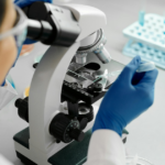 A scientist wearing blue gloves and safety goggles uses a pipette to place a sample on a glass slide under a microscope, with test tubes in a rack visible in the background.