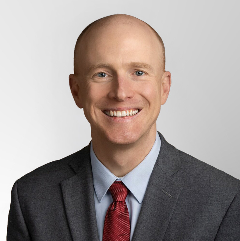 A smiling man with a bald head wearing a gray suit, light blue shirt, and red tie, posed in front of a plain light gray background.