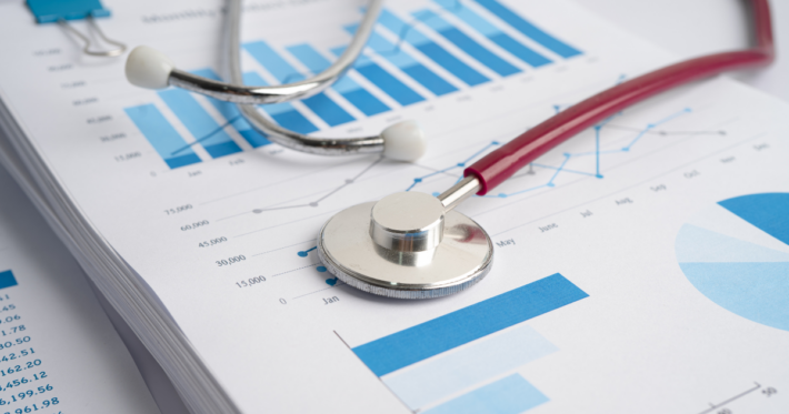 A stethoscope rests on printed charts and graphs with blue bars and lines, representing medical or healthcare statistics and data analysis.