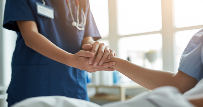 A healthcare professional in blue scrubs holds the hand of a patient in bed, offering comfort and support in a bright, sunlit room.