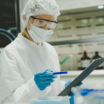 A scientist wearing protective gear, including a lab coat, mask, hair cover, and safety goggles, holds a pen and checks a clipboard while working in a laboratory setting with scientific equipment on the table.