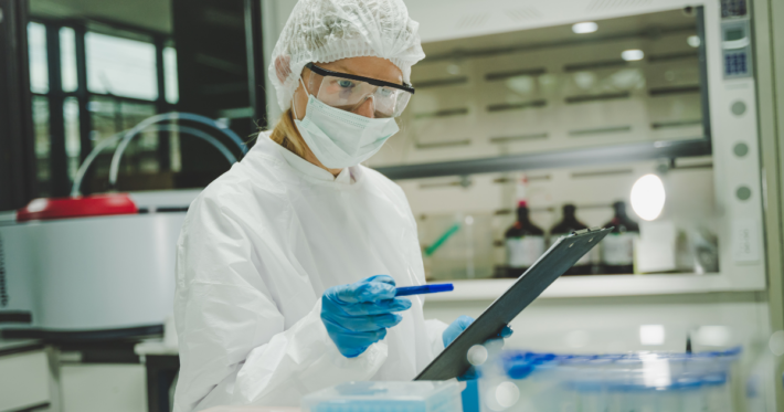 A scientist wearing protective gear, including a lab coat, mask, hair cover, and safety goggles, holds a pen and checks a clipboard while working in a laboratory setting with scientific equipment on the table.