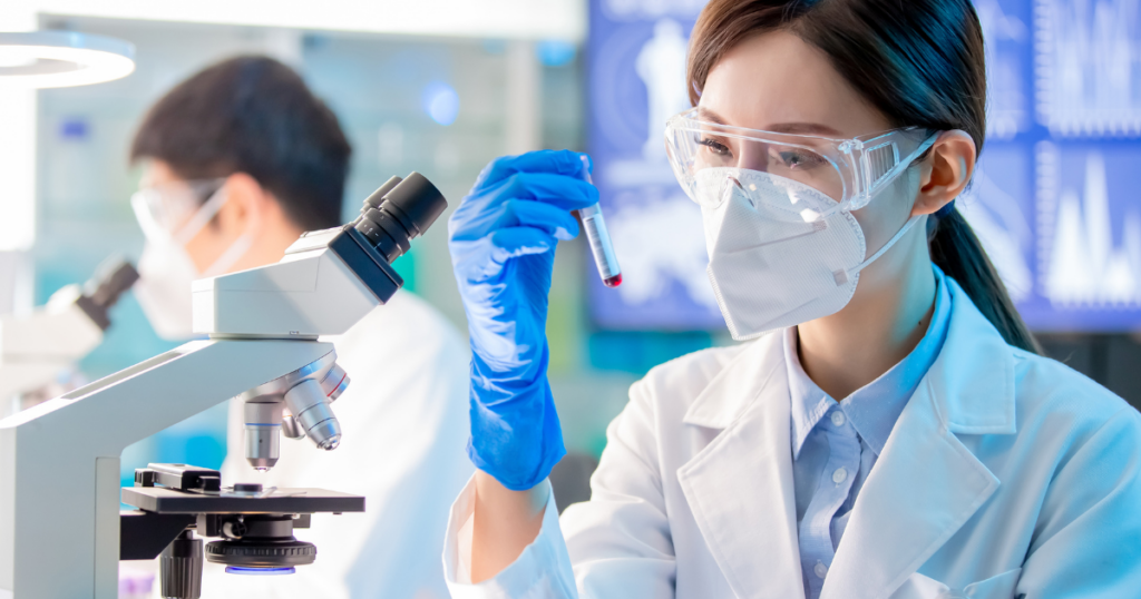 A scientist wearing a lab coat, gloves, safety goggles, and a mask examines a test tube beside a microscope in a laboratory. Another scientist works in the background. Scientific equipment and screens are visible.