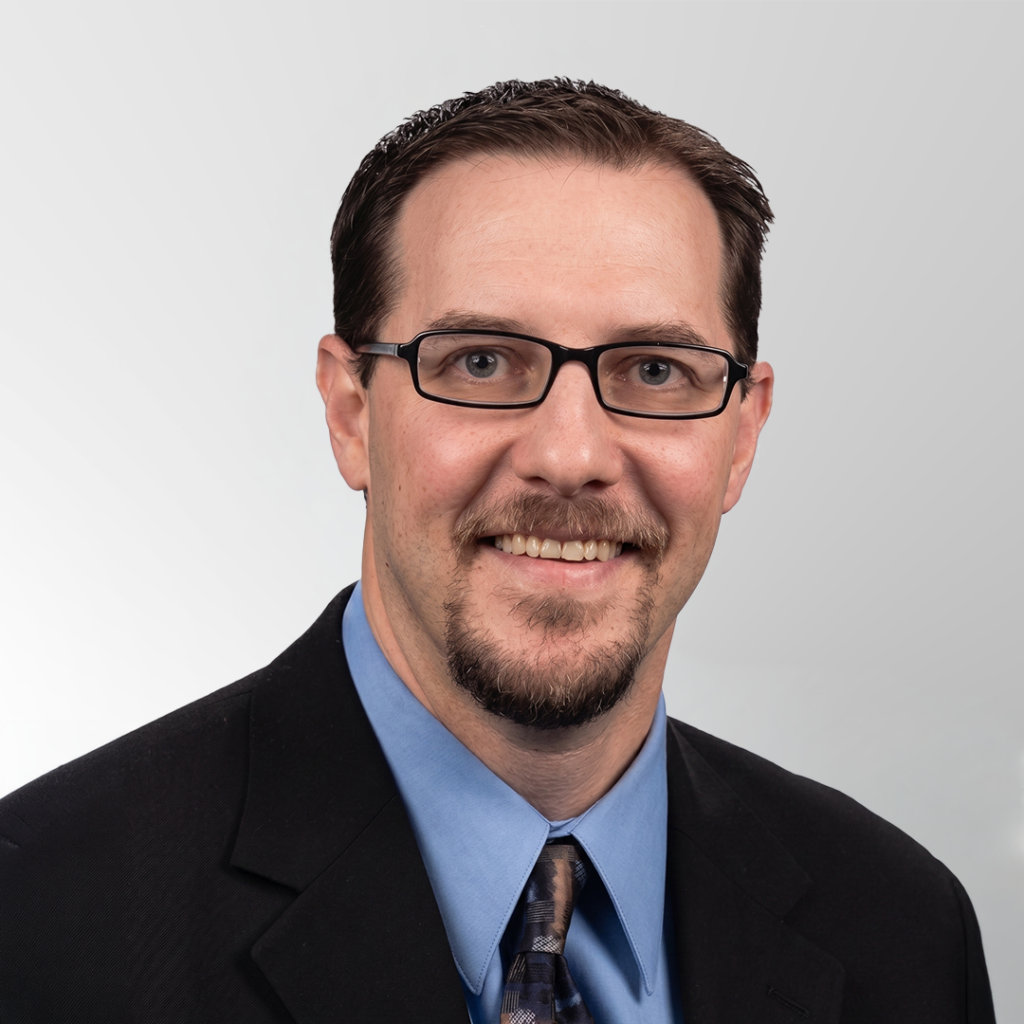 A man with short brown hair, glasses, and a goatee, wearing a black suit, light blue shirt, and patterned tie, smiling at the camera against a light gray background.
