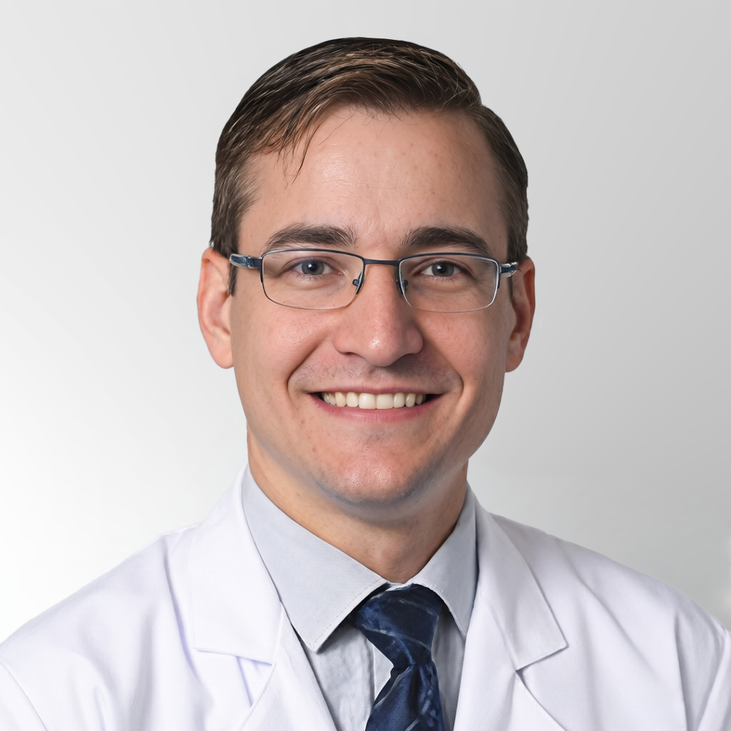 A young man with short brown hair and glasses, wearing a white lab coat, light blue shirt, and dark blue tie, smiles at the camera against a plain light gray background.