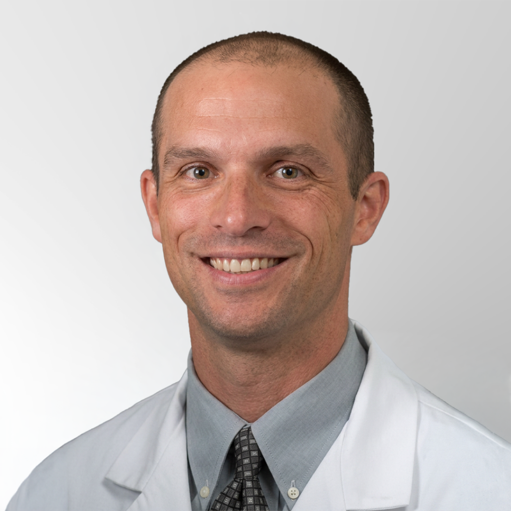 A smiling man wearing a white lab coat, gray dress shirt, and patterned tie, posing against a plain light gray background.