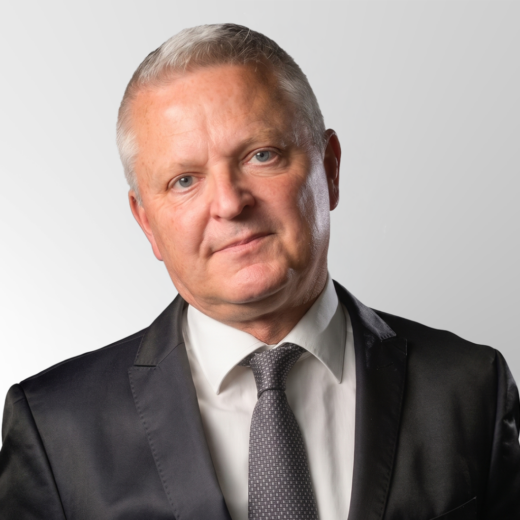 A middle-aged man with short gray hair wearing a black suit, white shirt, and gray patterned tie, standing against a plain light gray background, looking directly at the camera with a neutral expression.