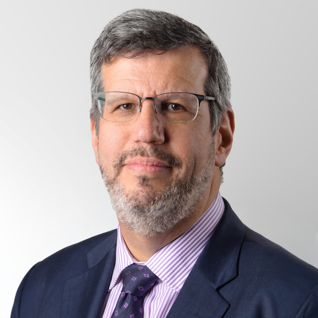 A middle-aged man with gray hair, glasses, and a beard, wearing a dark suit, light purple striped shirt, and a purple patterned tie, poses in front of a plain light background.