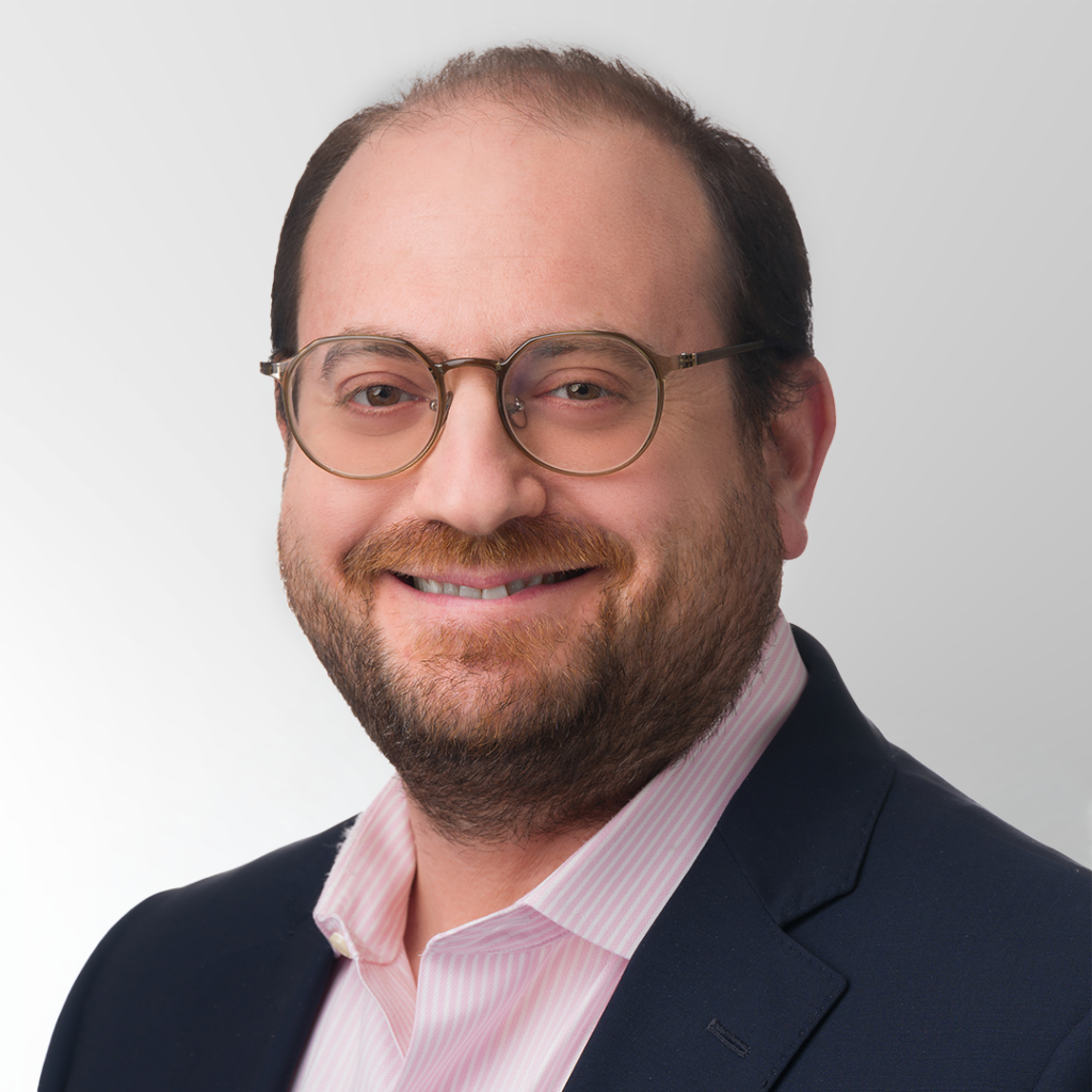 A smiling man with short brown hair, glasses, and a beard, wearing a dark suit jacket over a light pink collared shirt, poses in front of a plain, light gray background.