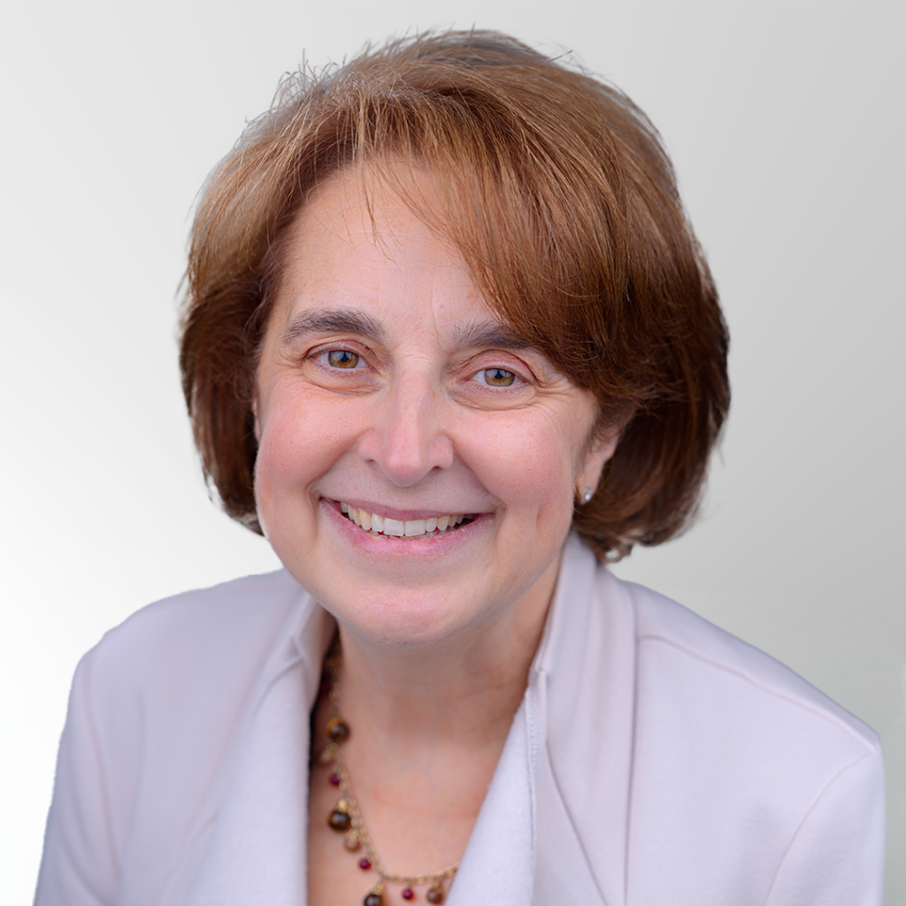 A middle-aged woman with short brown hair smiles at the camera, wearing a light-colored blazer and a beaded necklace, set against a light gray background.