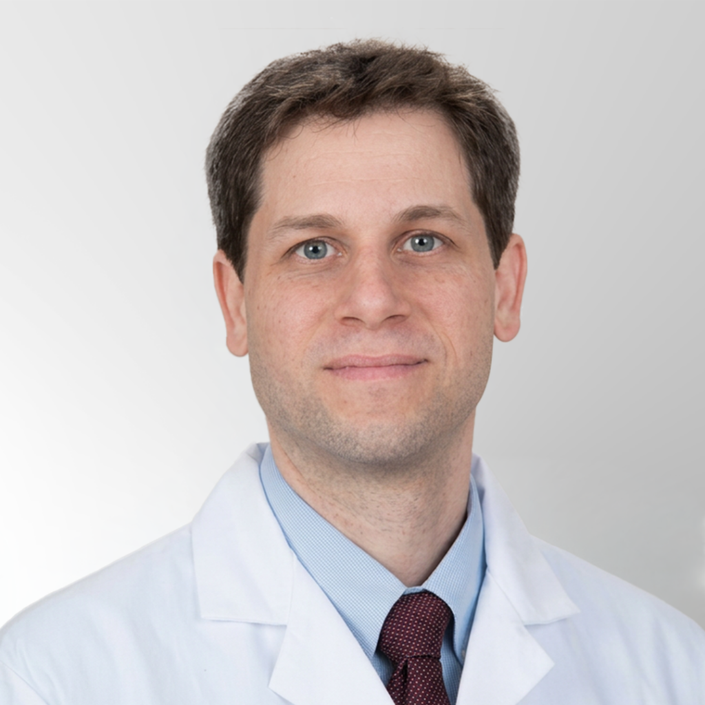 A man with short brown hair wearing a white lab coat, light blue shirt, and maroon tie, standing in front of a light gray background, looking directly at the camera.