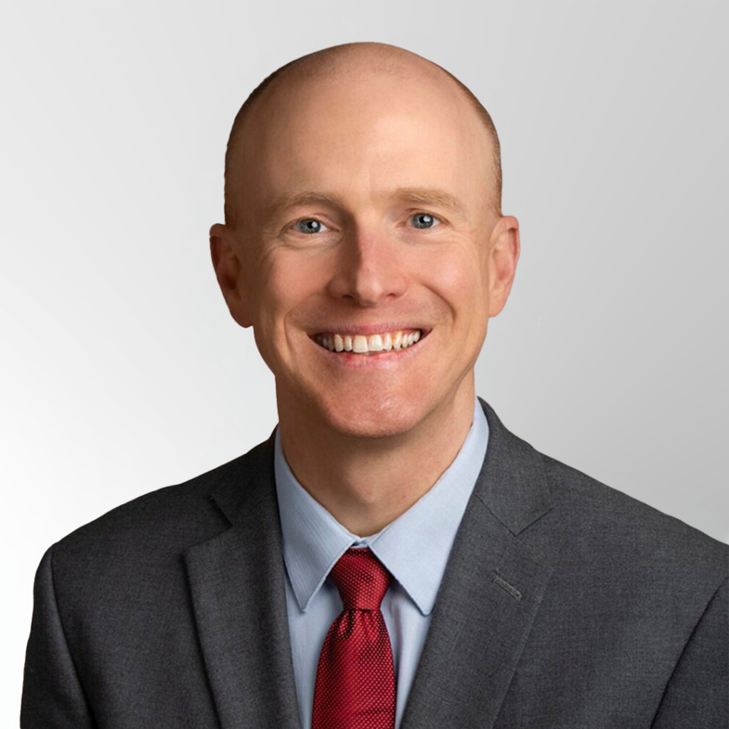 A smiling, bald man wearing a gray suit, blue shirt, and red tie, posed against a light gray background.