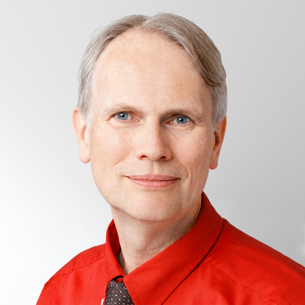 A middle-aged man with light skin, short gray hair, and blue eyes, wearing a bright red collared shirt and a dark patterned tie, smiles gently at the camera against a plain light background.