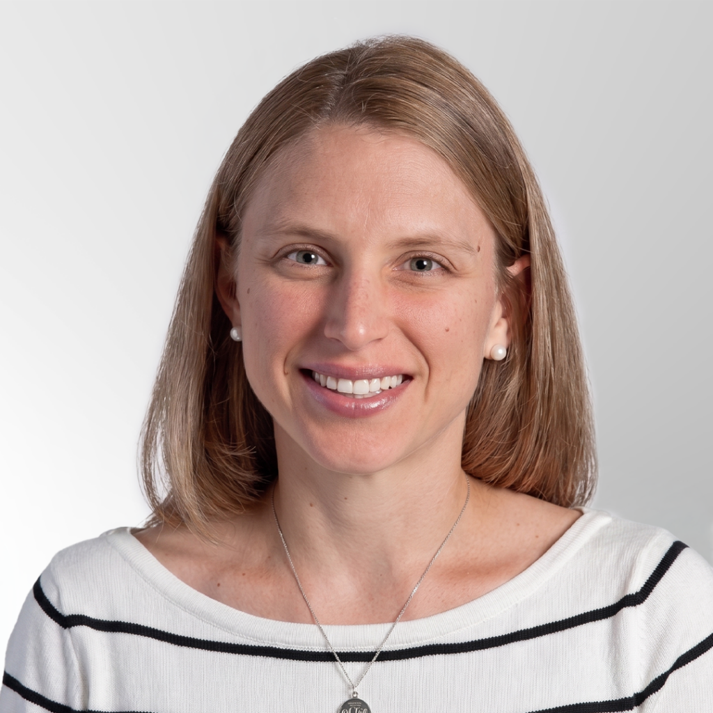 A woman with straight, shoulder-length light brown hair smiles at the camera. She is wearing a white top with black stripes, pearl earrings, and a silver necklace. The background is light gray.