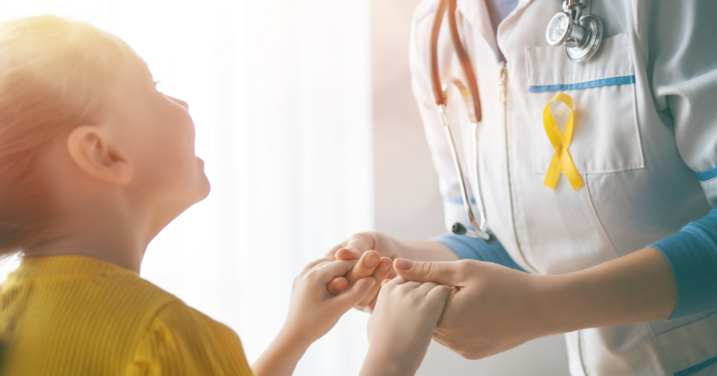 A doctor wearing a stethoscope and a yellow ribbon holds hands with a smiling child dressed in yellow, symbolizing support and care in a bright, sunlit room.