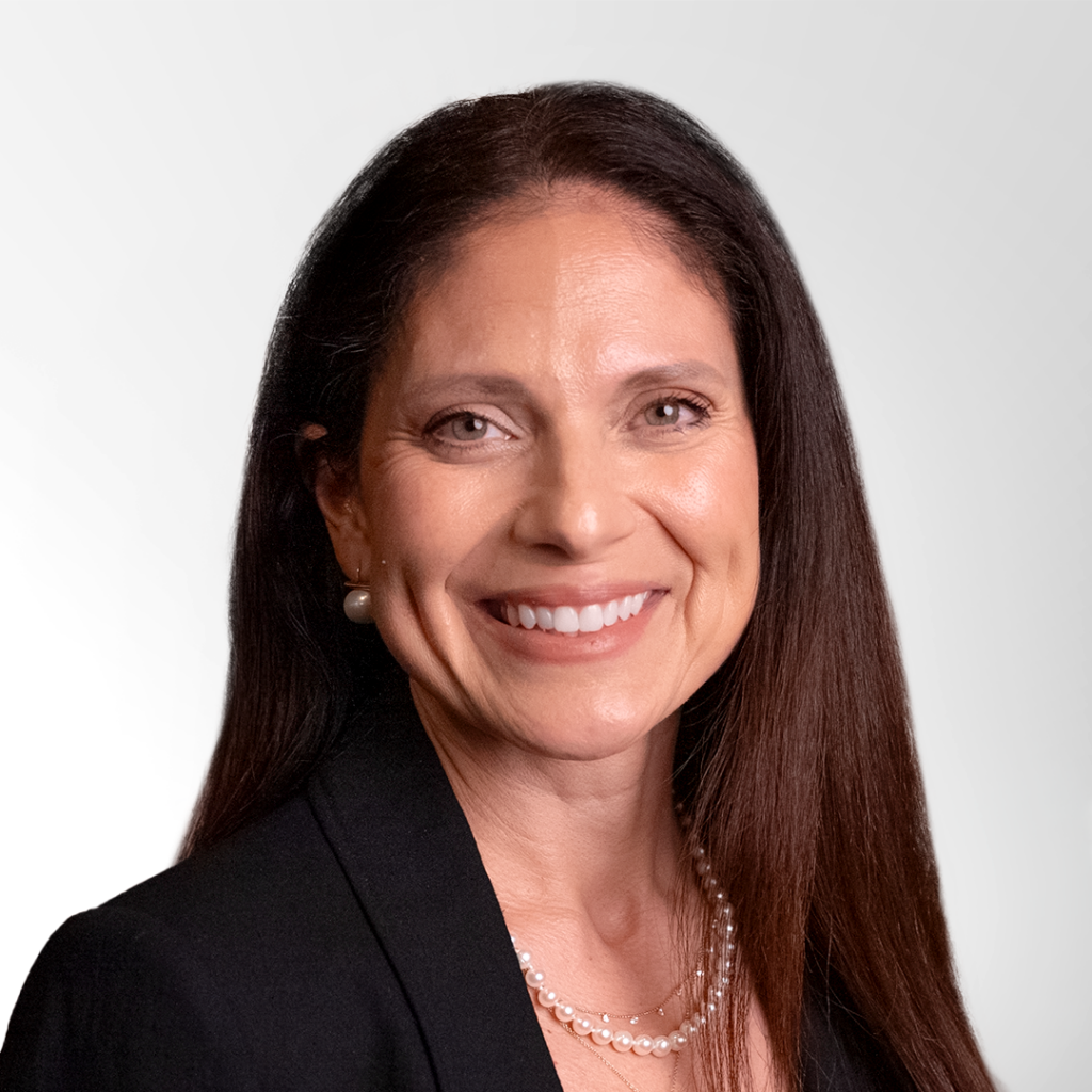 Smiling woman with long, straight brown hair wearing a black blazer, pearl necklace, and pearl earrings, posed in front of a light gray background.