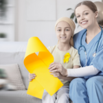A nurse and a young girl with a headscarf sit on a couch, smiling and holding a large yellow ribbon, symbolizing childhood cancer awareness.