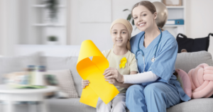 A nurse and a young girl with a headscarf sit on a couch, smiling and holding a large yellow ribbon, symbolizing childhood cancer awareness.