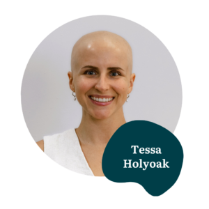 A smiling person with a bald head, wearing a white top and small hoop earrings. The name “Tessa Holyoak” is displayed in white text on a dark green abstract shape.
