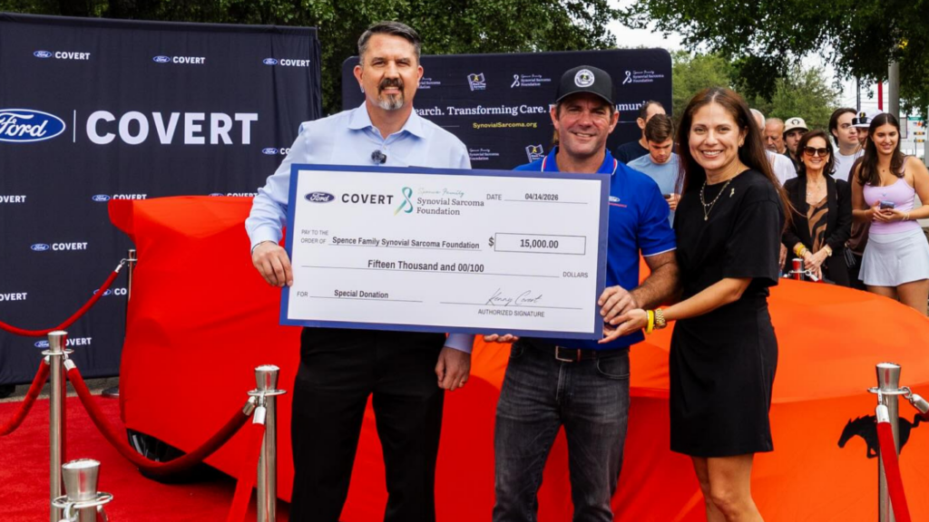 Three people stand on a red carpet holding an oversized $15,000 check from Covert Ford for a cancer foundation, with a covered car and a crowd in the background.