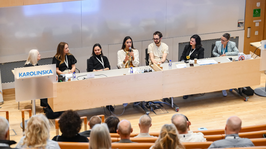 Six panelists sit at a long desk, speaking to an audience in a lecture hall. One panelist holds a microphone, while another sits at a podium labeled “Karolinska.” Bottles and papers are on the desk.