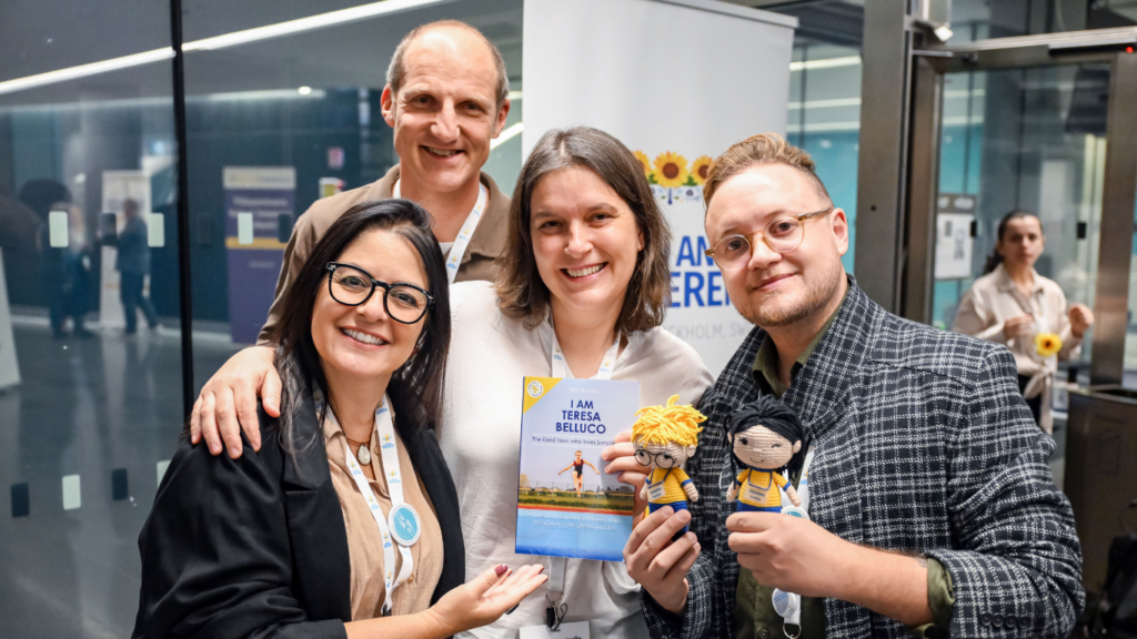 Four smiling adults pose indoors, holding a book titled “I Am Teresa Belluco” and two handmade dolls. They are standing close together in front of a glass door and informational posters.