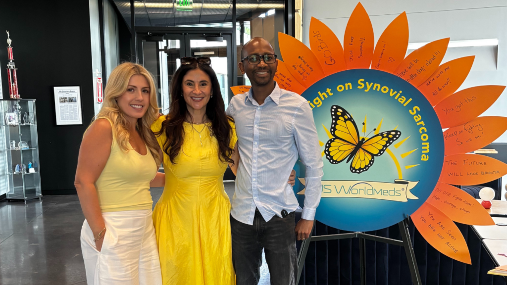 Three people stand smiling in front of a colorful sunflower-shaped sign that reads “Light on Synovial Sarcoma” with a butterfly in the center and handwritten messages on the petals.