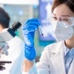 A scientist wearing a mask, goggles, and gloves examines a test tube in a laboratory. Another person works with a microscope in the background. Computer screens display data.