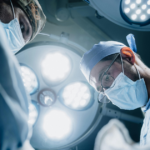Two surgeons in masks and scrubs focus on a patient under bright operating room lights, performing a surgical procedure.
