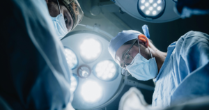 Two surgeons in masks and scrubs focus on a patient under bright operating room lights, performing a surgical procedure.