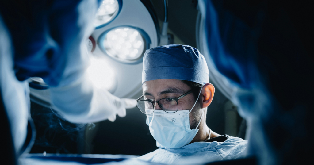 A surgeon wearing a mask, glasses, and surgical cap is focused during an operation. Bright surgical lights shine overhead, and another medical professional’s arm is visible in the foreground.