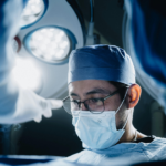 A surgeon wearing a mask, glasses, and surgical cap is focused during an operation. Bright surgical lights shine overhead, and another medical professional’s arm is visible in the foreground.