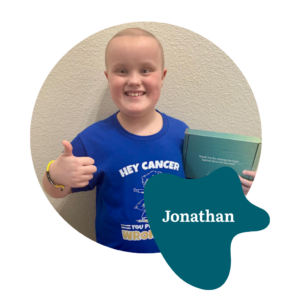A smiling child with a shaved head gives a thumbs up, wearing a blue “Hey Cancer, You Picked the Wrong Kid” shirt and holding a green box. The name “Jonathan” is displayed on a blue shape.