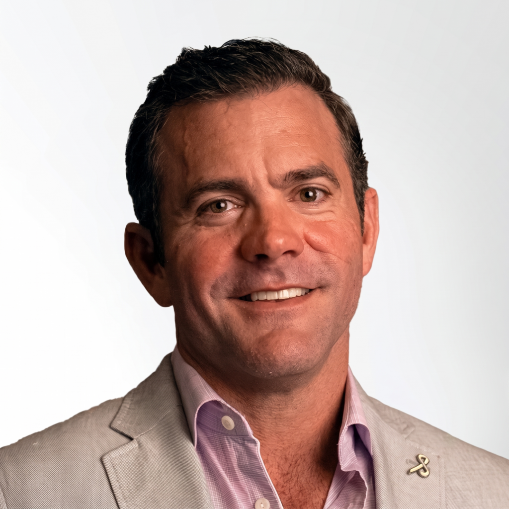 Man with short brown hair wearing a light gray blazer and a light purple shirt, smiling at the camera against a plain light background.