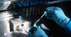 Hands wearing blue nitrile gloves draw liquid from a small glass vial into a syringe, with a rack of test tubes in the background.