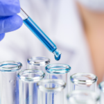 A gloved hand uses a dropper to add blue liquid into a row of clear glass test tubes in a laboratory setting.