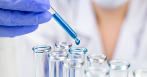 A gloved hand uses a dropper to add blue liquid into a row of clear glass test tubes in a laboratory setting.